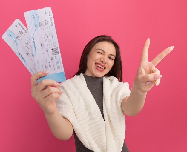 Joyful young pretty woman  stretching out airplane tickets  doing peace sign isolated on pink wall