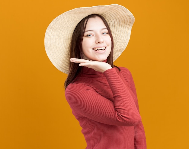 Joyful young pretty woman wearing beach hat standing in profile view looking at front keeping hand under chin isolated on orange wall with copy space