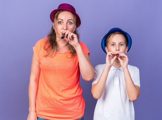 joyful young slavic boy with blue party hat standing with his mother wearing violet party hat blowing party whistle isolated on purple wall with copy space