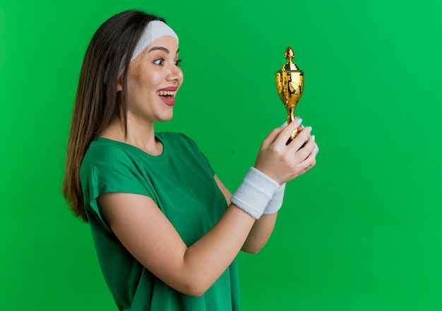 Free photo joyful young sporty woman wearing headband and wristbands standing in profile view holding and looking at winner cup