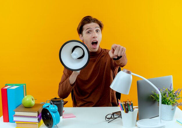 Joyful young student boy sitting at desk with school tools holding loudspeaker and showing you gesture 