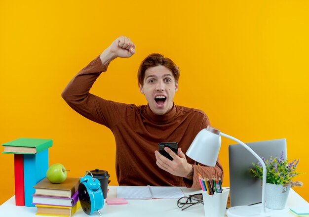 Joyful young student boy sitting at desk with school tools holding phone and raising hand 