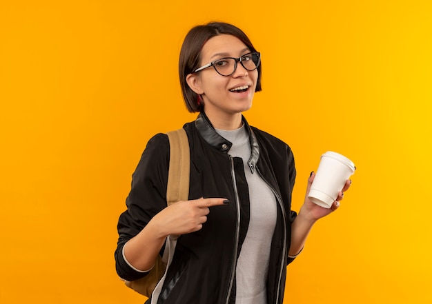 Joyful young student girl wearing glasses and back bag holding and pointing at plastic coffee cup isolated on orange