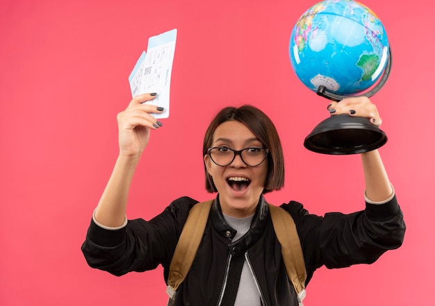 Free photo joyful young student girl wearing glasses and back bag raising airplane tickets and globe isolated on pink