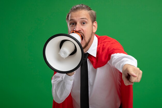 Joyful young superhero guy wearing tie speaks on loudspeaker and showing you gesture isolated on green background