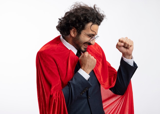 Joyful young superhero man in optical glasses wearing suit with red cloak stands sideways keeping fists isolated on white wall