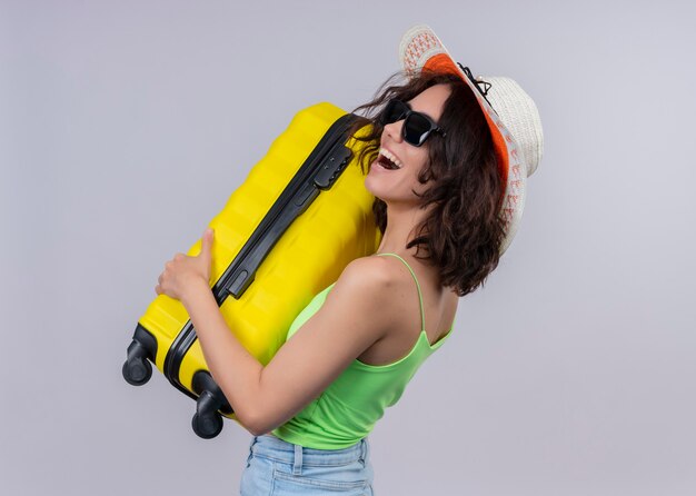 Joyful young traveler woman wearing hat and sunglasses and holding suitcase standing in profile view on isolated white wall