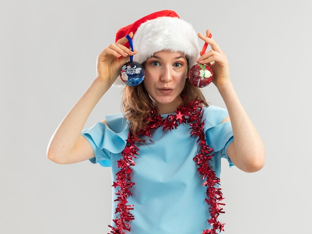 Free photo joyful young woman in blue top and santa hat with tinsel around her neck holding christmas balls smiling confident