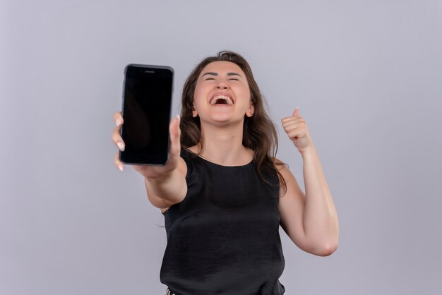 Joyful young woman wearing black undershirt held out phone to forward on white wall