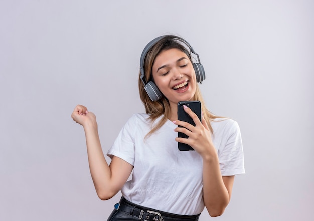 Free photo a joyful young woman in white t-shirt wearing headphones singing while listening to the music on her phone on a white wall