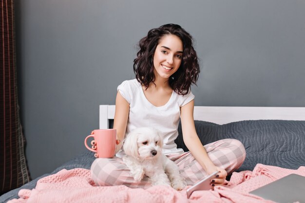 Joyful young woman with curly brunette hair in pajamas chilling on bed with little dog in modern appartment. Pretty model relaxing at home with cup of coffee, chatting on phone, smiling