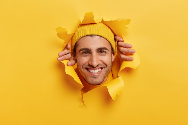 Free photo joyous caucasian man with toothy smile, shows white teeth, looks through hole torn in paper