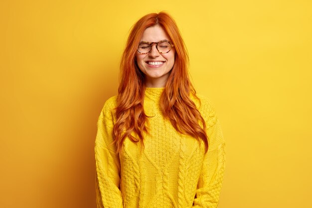 Joyous pretty woman with red hair closes eyes stands satisfied dressed in casual jumper wears optical glasses recalls somethig very pleasant.