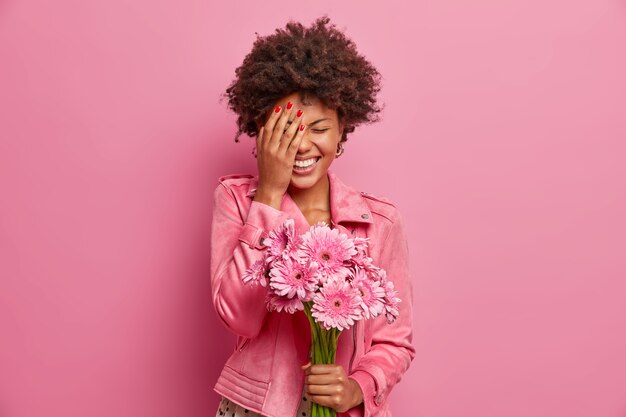 Joyous young African American woman laughs happily, makes face palm, gets nice gift like flowers, holds beautiful gerberas, expresses sincere emotions, 