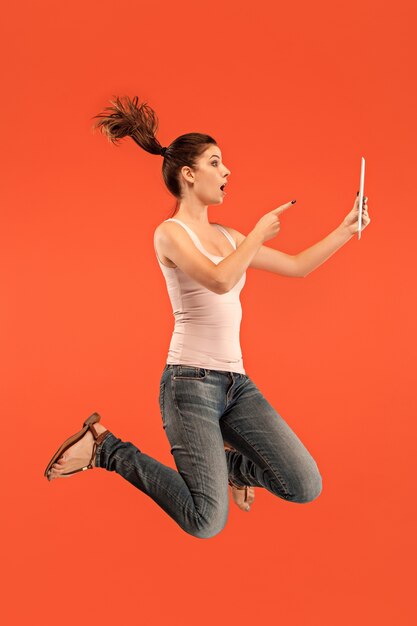 Jump of young woman over blue studio background using laptop or tablet gadget while jumping. Runnin girl in motion or movement. Human emotions and facial expressions concept. Gadget in modern life