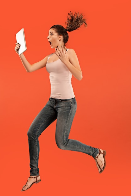 Jump of young woman over blue studio background using tablet gadget while jumping.