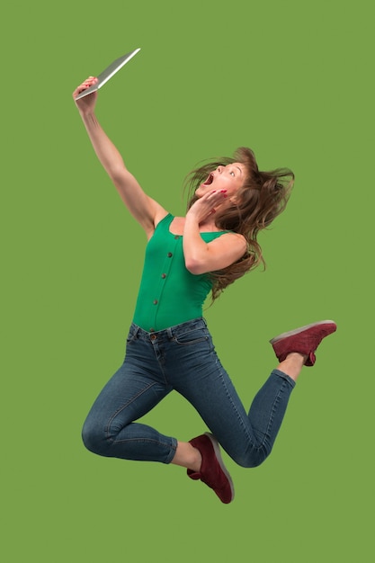 Free Photo jump of young woman over green studio background using laptop or tablet gadget while jumping. runnin girl in motion or movement. human emotions and facial expressions concept. gadget in modern life