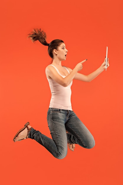 Jump of young woman over orange studio wall using tablet