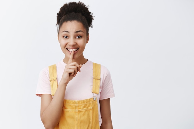 Keep secrets safe. Portrait of charming friendly-looking cute African American female in yellow trendy overalls, saying shh while showing shush gesture, smiling and holding index finger over mouth