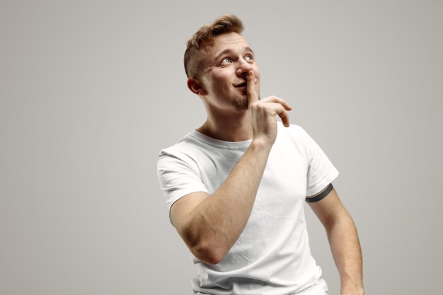 Keep silence. Handsome young man in white shirt looking at camera and holding finger on lips while standing against grey background