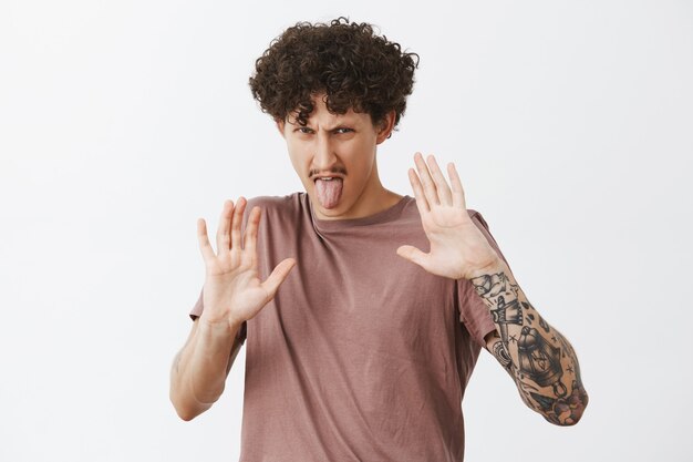 Keep this thing away from me. Stylish tattooed man expressing disgust and aversion raising palms to protect himself from food he dislikes sticking out tongue and frowning from displeased emotions