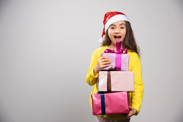 Kid dressed in Santa Claus red hat carrying a lot of Christmas presents. High quality photo