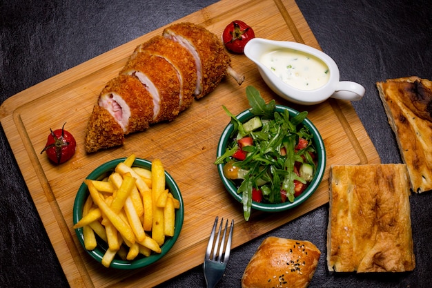 Kiev style cutlet on the wooden board with french fries arugula tomato cucumber sauce top view