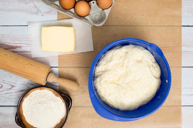 Kneaded dough in blue bowl; eggs; butter; flour; and rolling pin on wooden desk