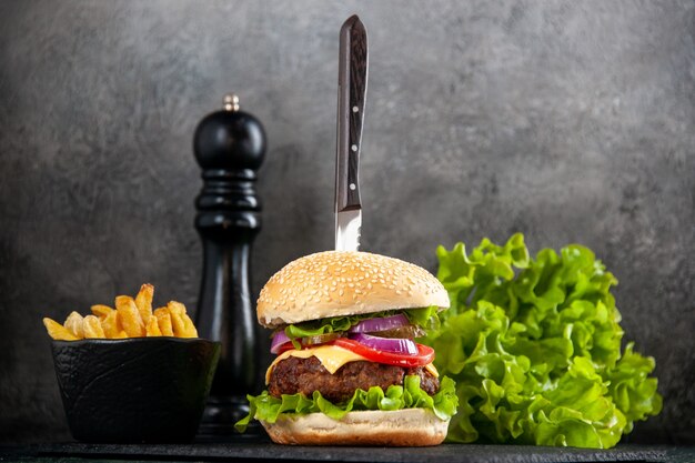 Knife in delicious meat sandwich and green fries on black tray on gray surface