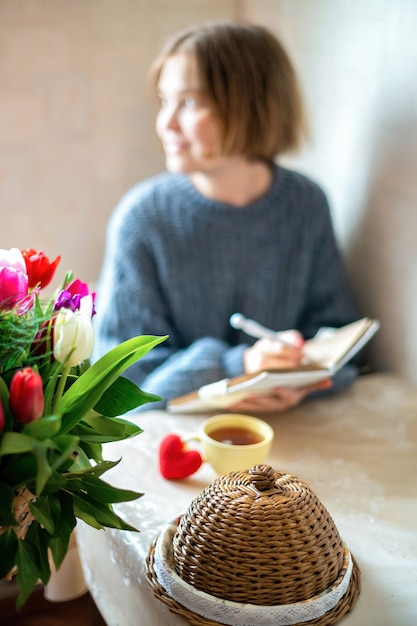 Free photo knitted red heart and a cup of tea on the table with a girl holding notebook. kitchen