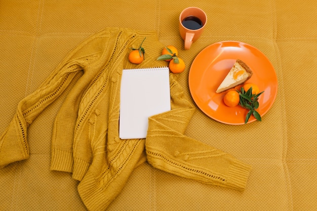 Free photo knitted sweater, cup of coffee, cheesecake, mandarines, notebook on orange table