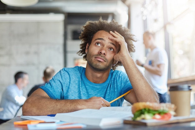 Knowledge and education. Unhappy Afro American university student wearing blue t-shirt looking up with questionable frustrated expression, feeling tired while working on home assignment at cafe