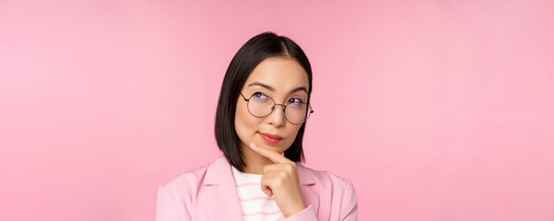 Korean businesswoman thinking wearing glasses looking thoughtful at camera making decision standing over pink background