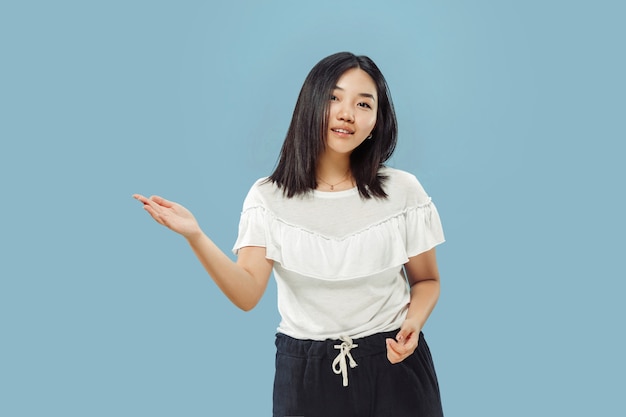 Korean young woman's half-length portrait on blue  space. Female model in white shirt. Showing and pointing something.