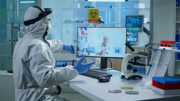 Free photo lab technician in ppe suit sitting at table in medical research laboratory, holding test tube and talking on video call with senior doctor giving online advices during virtual meeting