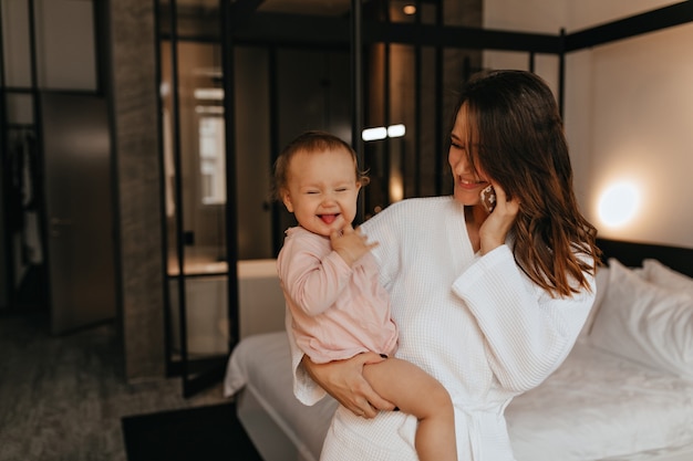 Lady in bathrobe in excellent mood is talking on phone, sitting on bed with perky female baby showing her tongue.