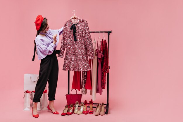 Lady in blouse with flounces and red beret is looking at elegant shiny pink dress.