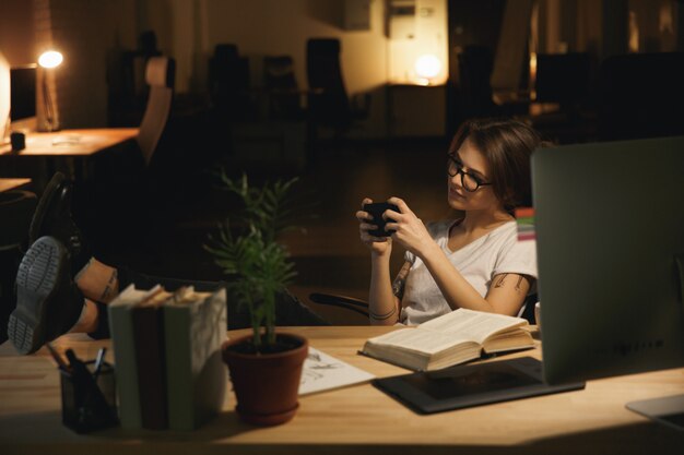 Lady designer sitting indoors at night play games by phone.