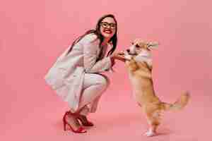 Free photo lady in eyeglasses and suit plays with corgi on pink background.  happy woman in office attire and red high heels smiles and holds corgi.