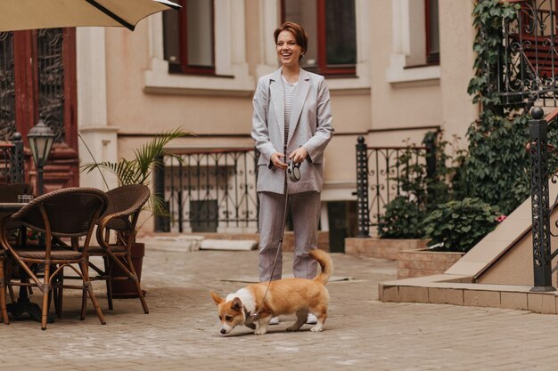 Lady in grey suit walks with her corgi. Portrait of happy woman in jacket and pants posing with dog outside