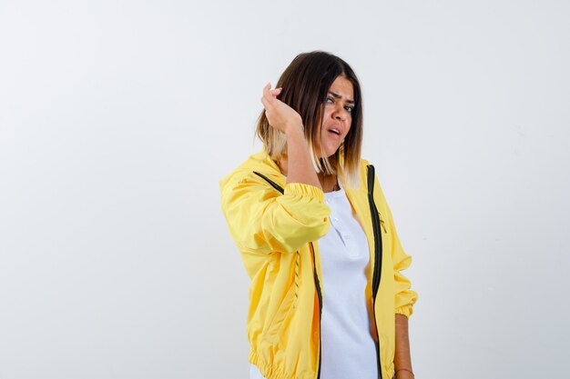 Lady keeping hand behind ear in t-shirt, jacket and looking confused. front view.