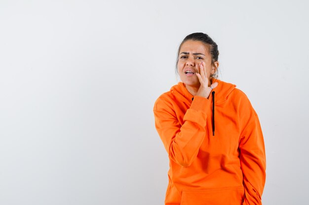 Lady in orange hoodie telling secret behind hand and looking concerned, front view.