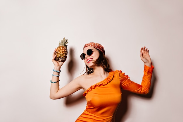 Lady in orange outfit laughs, dances and holds pineapple on white space.