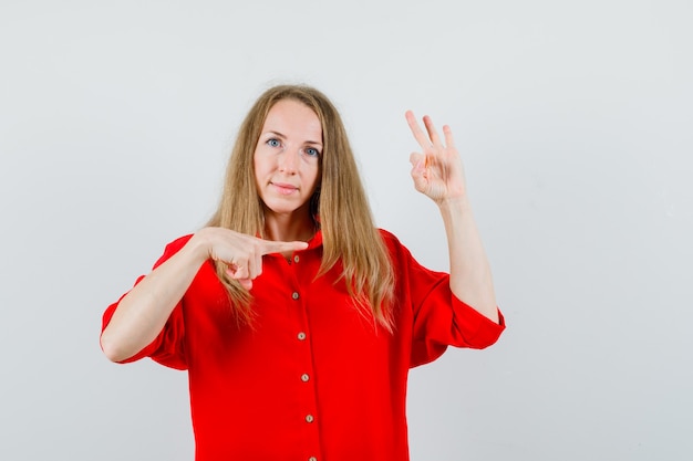 Lady pointing aside, showing ok sign in red shirt and looking confident.