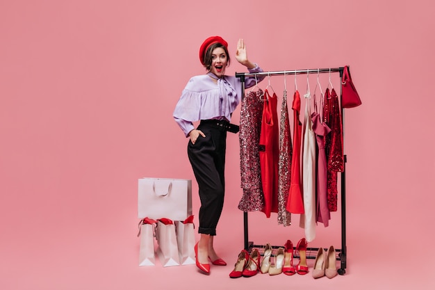 Free Photo lady poses in dressing room with bright clothes and shoes. girl in beret and lilac blouse looking at camera on pink background.