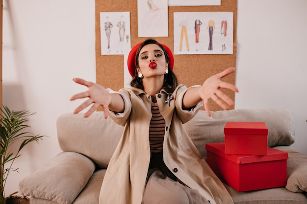 Free photo lady in red beret and beige outfit blows kiss. cute young woman with red lips in long fashionable coat posing at camera.