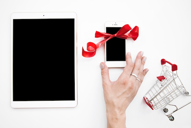 Free photo lady's hand on smartphone near tablet and shopping trolley