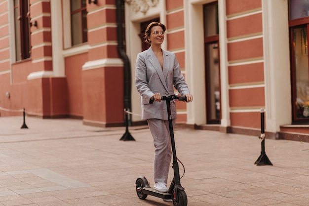 Lady in stylish suit having fun and riding scooter. Pretty happy woman in grey oversize jacket and pants smiling and enjoying city view