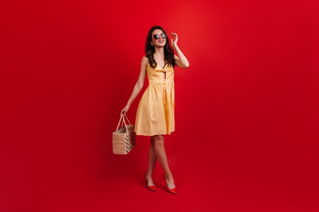 Lady in stylish summer outfit posing on red background Curly brunette in sunglasses holds beach bag