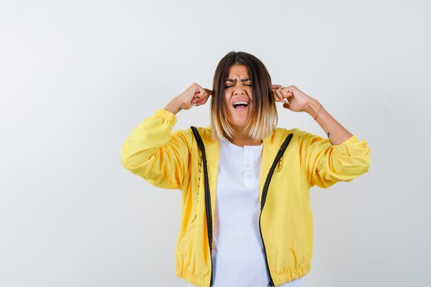 Lady in t-shirt, jacket plugging ears with fingers and looking annoyed , front view.